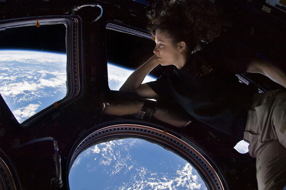 A casually dressed astronaut rests against the round earth-filled windows of the International Space Station's Cupola module.