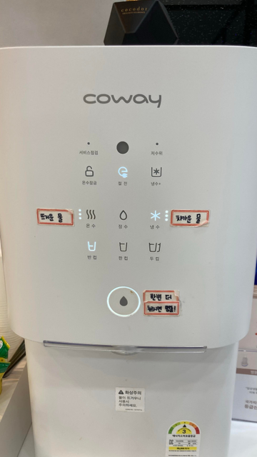Coway 정수기 정면. 하얀색 바탕에 회색 아이콘, 그 밑에 좀 더 진한 회색으로 작은 글씨가 써있는 구성. 위에서부터 서비스점검, 저수위(이상 아이콘 없음), 온수잠금, 절전, 냉수, 온수, 절수, 냉수, 한 잔, 한 컵, 두 컵, 그리고 글씨 없이 물방울 그림만 있는 커다란 동그란 버튼.
그리고 이들 버튼 옆에 견출지가 붙어있다. 굵은 손글씨로 각각 ‘뜨거운 물’(온수 옆에), ‘차가운 물’(냉수 옆에), 그리고 ‘한번 더 누르면 멈춤!’(동그란 버튼 옆에)이라고 써있다.