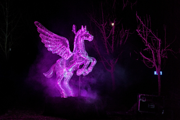 A night-time photo of a rearing disco pegasus illuminated with purple lights.
Yes, you read/heard right, a disco pegasus. A pegasus (horse with wings), covered in tiny mirrors, like a disco ball.
There seems to be a fog machine nearby, because some purple (because of the lights) fog seems to be rising from the ground around the pegasus, giving the whole image a majestic vibe (besides the fact that it is a purple disco pegasus, of course).
Otherwise, there are a few trees illuminated in purple in the background as well. And a box of some sort.
But mainly: Purple Disco Pegasus.