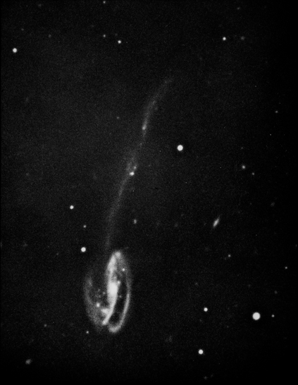 A grainy, black and white image of a galaxy. In the upper left corner is a spiral galaxy with a bright central core encircled by two wispy arms that form a loose spiral. The view is somewhat oblique, giving the galaxy an oval appearance. A long, thin, diffuse stream extends from the lower right side of the galaxy down to the lower right corner of the image. In the background there are a few circular dots, which are stars, and smudgy objects, which may be background galaxies.