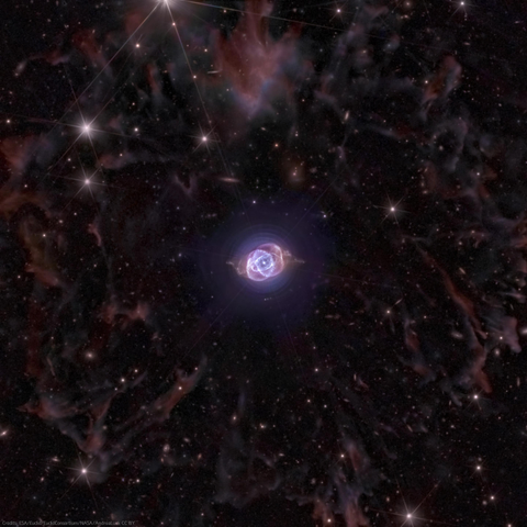 This image presents a stunning deep space view centered on the Cat’s Eye Nebula, with intricate structures and vibrant colors. The nebula itself glows in an ethereal blue and violet hue, surrounded by delicate, symmetrical, and concentric gaseous layers that form its iconic shape. The surrounding space is filled with an intricate web of faint reddish and brownish wisps on the outer region surrounding the nebula. Bright stars with distinct diffraction spikes punctuate the scene, adding depth and contrast to the dark, star speckled background. Numerous galaxies are also visible in the background. The image showcases a great balance between sharp details in the nebula's core and the softer, ghostly tendrils of interstellar gas beyond.