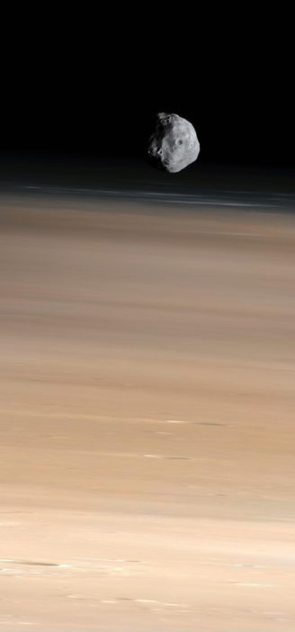 An image taken from orbit shows the potato-shaped satellite Phobos orbiting Mars. The perspective places Phobos very close to the Martian limb, above elongated whitish clouds near the terminator. Phobos appears predominantly grey, while the surface of Mars displays tones ranging from rusty red to darker hues near its terminator. Some craters are visible on the surface of Mars, they should be much larger than Phobos. The camera scanning the satellite causes the view of the Martian surface to appear stretched as it moves by.