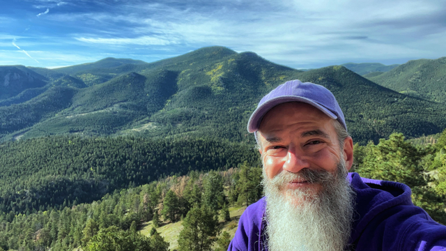 Selfie of a smiling man with a gray mustache and long, gray beard, wearing a purple hoodie and a purple baseball cap, perched on the edge of a rock in a Colorado state park with beautiful rolling hills and mountains covered in dark green pine trees stretching off into the distance behind, with mostly sunny, blue skies above, just a few, wispy cirrus clouds above the hills.