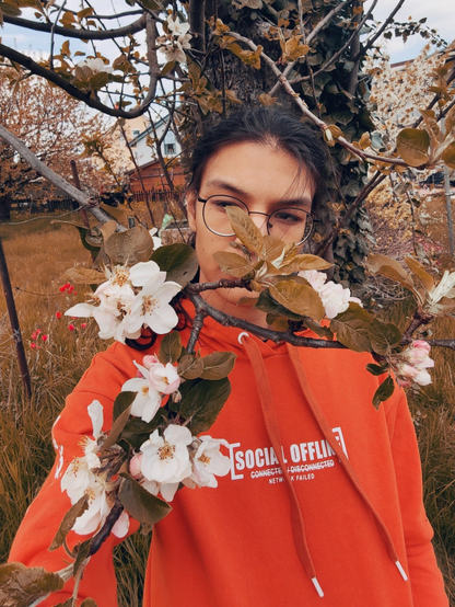 Selfie, looking sideways. Apple tree with ivy in the background and some twigs and blossoms in front of my face. Wearing an orange hoodie dress that says: “Social offline / Network failed”