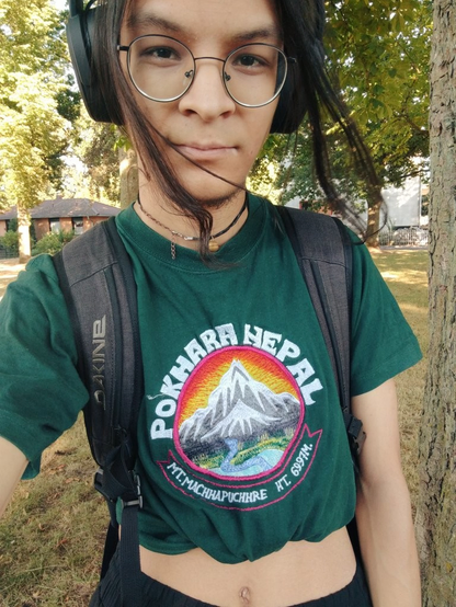 Selfie, outside, sunny. Wearing headphones. Strands of hair waving in the wind. Shirt is tied up/cropped, says: “Pokhara Nepal” with colorful embroidery of mountains and valley.