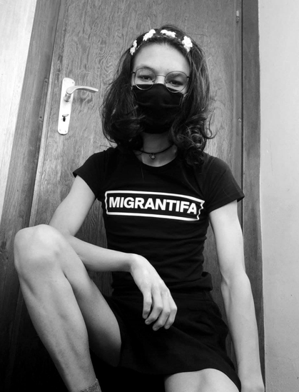 Selfie in black and white, kinda crouching, one arm sitting on my leg. Wearing a face mask in black, choker, hair band with flowers on top, a black skirt and my black shirt says “Migrantifa”.